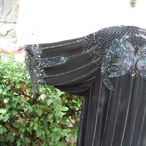 May include: Black evening gown with iridescent sequin and bead embellishments. The gown features a unique design on the shoulders and sleeves, with vertical lines down the front. The sleeves have fringe detailing. The garment is made of a flowing, lightweight fabric.