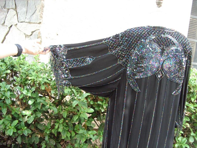 May include: Black evening gown with iridescent sequin and bead embellishments. The gown features a unique design on the shoulders and sleeves, with vertical lines down the front. The sleeves have fringe detailing. The garment is made of a flowing, lightweight fabric.
