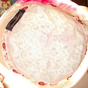 May include: A cream-colored hat with a floral patterned lining and a white lace interior. The hat has a pink floral trim and a black label that reads "Suzanne Custom Designed". The hat is made of a soft, satin-like material.