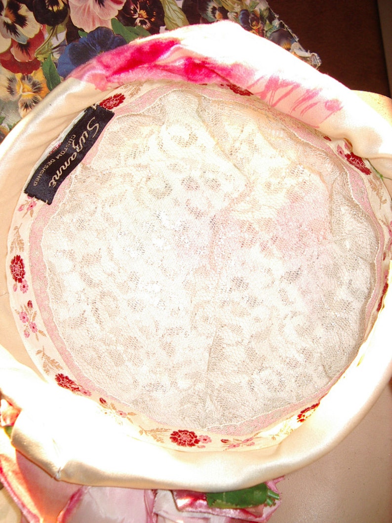 May include: A cream-colored hat with a floral patterned lining and a white lace interior. The hat has a pink floral trim and a black label that reads "Suzanne Custom Designed". The hat is made of a soft, satin-like material.