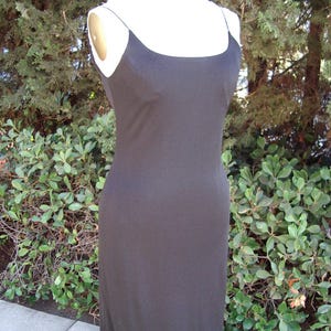 May include: A black, floor-length dress with thin spaghetti straps. The dress is displayed on a mannequin against a backdrop of green foliage. The dress has a simple, elegant design.