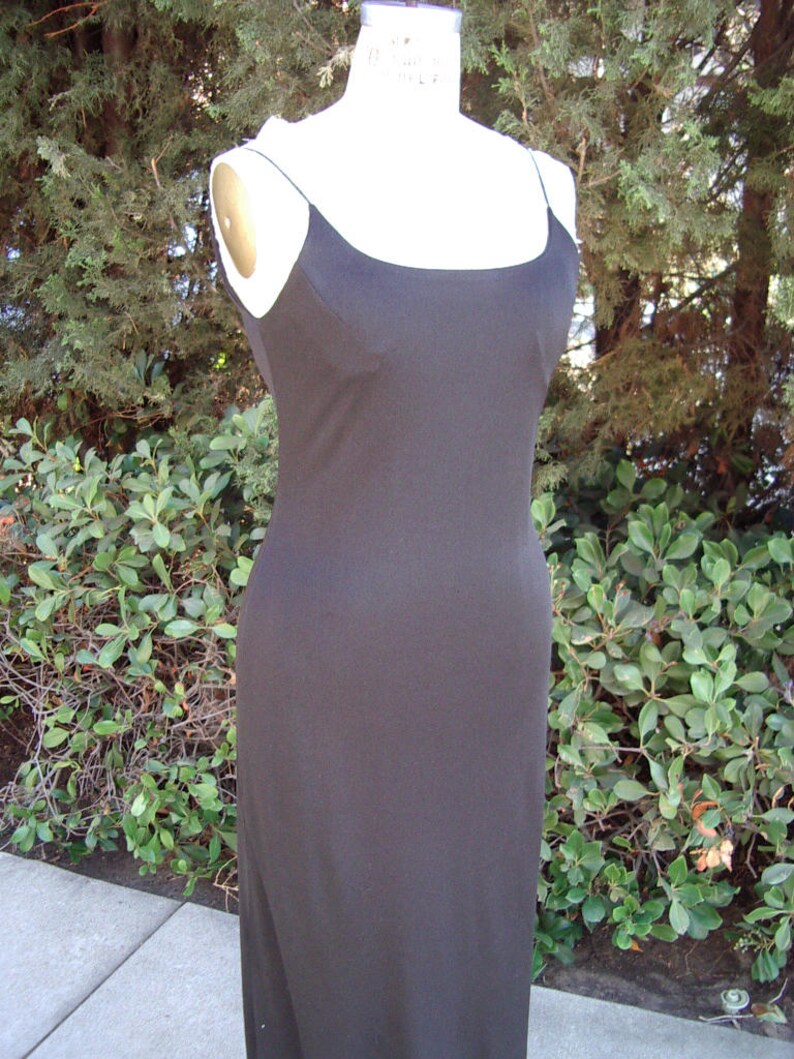 May include: A black, floor-length dress with thin spaghetti straps. The dress is displayed on a mannequin against a backdrop of green foliage. The dress has a simple, elegant design.
