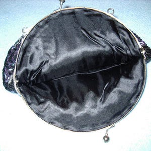 May include: Open view of a vintage black beaded handbag with a silver-toned frame. The interior is lined with black satin fabric. The bag's exterior features a beaded design with hints of purple and silver.