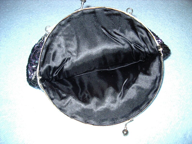 May include: Open view of a vintage black beaded handbag with a silver-toned frame. The interior is lined with black satin fabric. The bag's exterior features a beaded design with hints of purple and silver.
