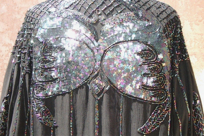 May include: Black evening top with iridescent sequin and bead embellishments. The top features a detailed design on the bodice and sleeves, with a square neckline and a flowing, sheer fabric. The sequins shimmer with multiple colors.