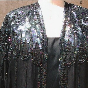 May include: Black evening jacket with iridescent sequin and bead embellishments. The jacket features a shawl collar and long sleeves. The sequins and beads are concentrated on the shoulders and along the edges, creating a shimmering effect. The jacket is worn over a black dress.