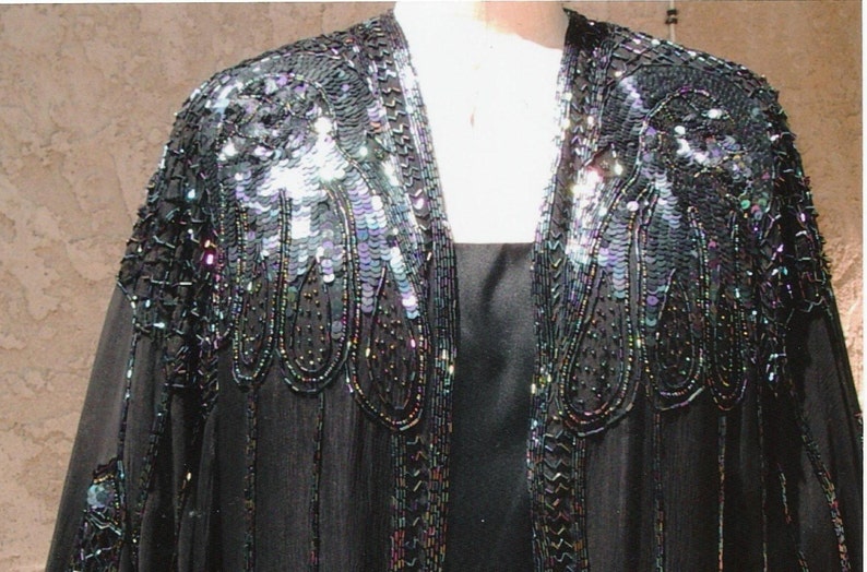 May include: Black evening jacket with iridescent sequin and bead embellishments. The jacket features a shawl collar and long sleeves. The sequins and beads are concentrated on the shoulders and along the edges, creating a shimmering effect. The jacket is worn over a black dress.