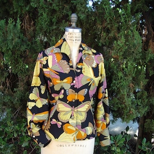 May include: A black button-down shirt with a vibrant butterfly pattern in yellow, orange, pink, and brown. The long-sleeved shirt is displayed on a mannequin, showcasing its collared neckline and buttoned cuffs. The background is a lush green bush.