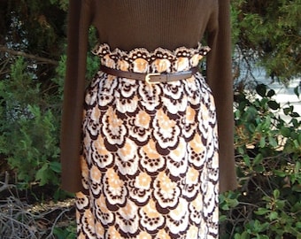 May include: A brown turtleneck sweater paired with a patterned skirt. The skirt features a repeating design in white, brown, and orange. A brown belt cinches the waist. The outfit is displayed on a mannequin.