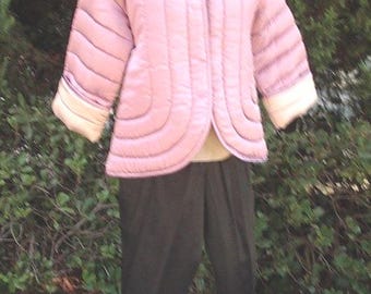 May include: A mauve quilted jacket with a V-neckline, three-quarter length sleeves, and light-colored cuffs, paired with solid black trousers. The jacket features horizontal stitched channels throughout the body and sleeves, worn on a mannequin.