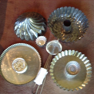 May include: A set of six silver-toned metal baking molds. The molds are in various shapes and sizes, including a round mold with a fluted edge, a round mold with a center hole, and a small, round mold with a fluted edge. The molds are all made of a shiny metal and have a smooth surface.