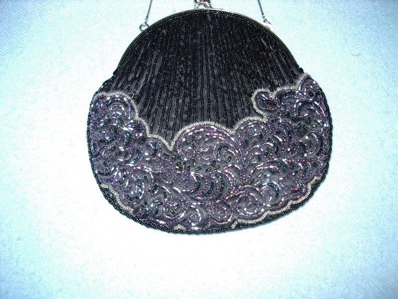 May include: A black beaded evening bag with a silver chain handle. The bag has a rounded shape with a scalloped edge. The bottom edge features a decorative, swirling pattern in shades of purple and silver.