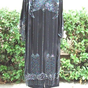 May include: Black, long-sleeved dress with intricate sequin and bead embellishments. The dress features a detailed design on the shoulders and lower hem, with vertical beaded lines and fringe. The dress is displayed on a rolling stand.