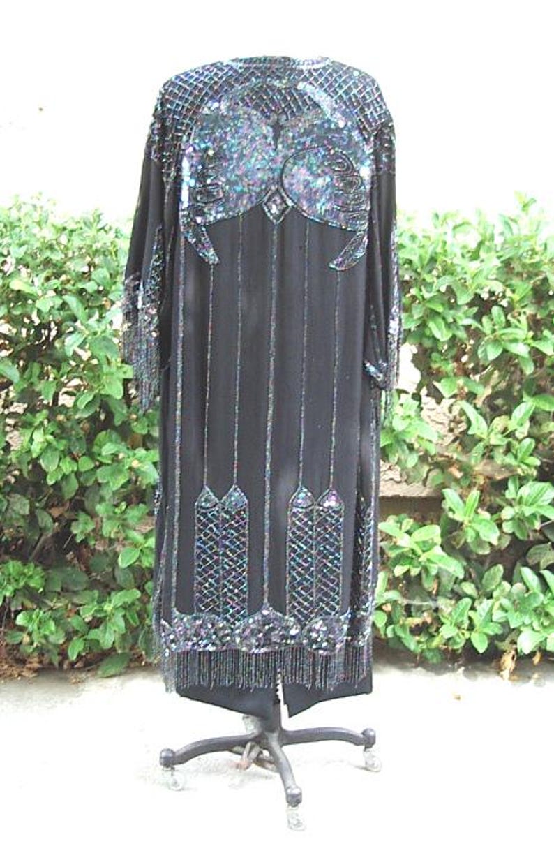 May include: Black, long-sleeved dress with intricate sequin and bead embellishments. The dress features a detailed design on the shoulders and lower hem, with vertical beaded lines and fringe. The dress is displayed on a rolling stand.