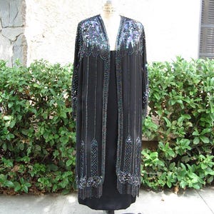 May include: A black, floor-length dress with a matching beaded jacket. The jacket features intricate sequin and bead detailing in shades of blue, green, and silver along the neckline, sleeves, and front edges. The dress is displayed on a vintage dress form.