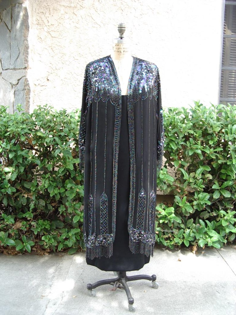 May include: A black, floor-length dress with a matching beaded jacket. The jacket features intricate sequin and bead detailing in shades of blue, green, and silver along the neckline, sleeves, and front edges. The dress is displayed on a vintage dress form.