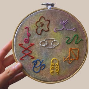 May include: A circular embroidery hoop with a colourful, abstract design. The design features various shapes and lines in red, pink, orange, yellow, green, blue, and purple thread. The fabric has a watercolour-like effect.