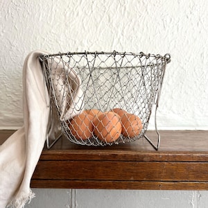 May include: A silver-toned wire mesh basket with a tapered shape and a metal frame, containing three brown eggs. A light beige fabric cloth with a frayed edge is draped over the left side of the basket, partially covering the wire structure.