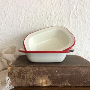 May include: Two white enamel bowls with a red rim are stacked on a vintage book. The bowls have a rectangular shape with rounded corners. The background is a textured white wall.