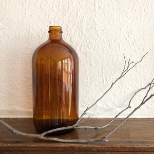 May include: A large, amber glass bottle with a rounded top and vertical ridges. The bottle is on a wooden surface, with bare branches in the foreground. The background is a textured white wall.