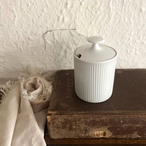 May include: A white ceramic sugar bowl with a lid and a small spoon. The bowl has vertical ribbed detailing and a round handle on the lid. The bowl is on a vintage book.