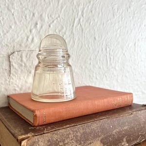 May include: A clear glass electrical insulator with a tiered, dome-shaped top and a wide base. The side of the glass insulator features the embossed text "HEMINGRAY" and "NO 9". The object is positioned on top of a closed, orange-coloured hardcover book.
