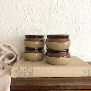 May include: Four small, round ceramic pots stacked on a vintage book. The pots are two-toned, with a light tan base and a dark brown upper section. The book has a beige cover and text on the spine.