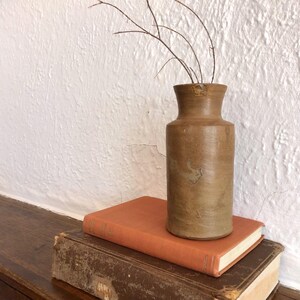 May include: A brown ceramic vase with a narrow neck and a cylindrical body, containing thin, dried branches. The vase is placed on two antique books, one with an orange cover and the other with a brown cover.
