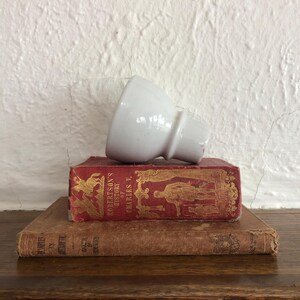May include: A white ceramic object, possibly a lamp part, sits on two antique books. The top book is red with gold lettering and illustrations. The bottom book is brown with faded text.
