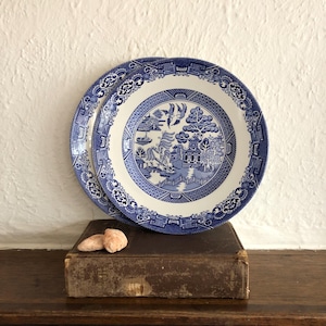 May include: Two blue and white ceramic plates featuring a detailed Willow pattern design. The plates are stacked on a vintage book, with two small, light-coloured stones on the cover. The plates have a classic, decorative style, perfect for a dining table.