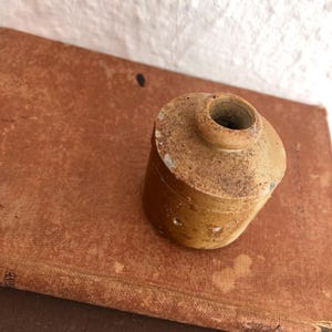 May include: A small, antique, cylindrical ceramic jar with a rounded top and a small opening. The jar is a mottled tan colour with darker brown spots and sits on a worn, reddish-brown surface. The jar appears to be aged.