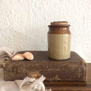 May include: A small, two-tone ceramic jar with a cork stopper rests on a vintage book. The jar is tan with a brown rim. Two small, light-coloured stones are next to the jar. The book is brown with worn edges.