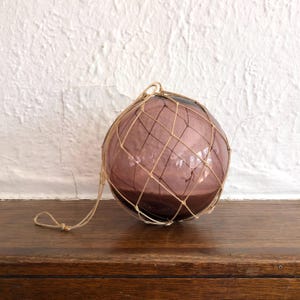 May include: A spherical, purple glass float, held within a beige net, sits on a wooden surface. A rope loop is attached for hanging. The background is a textured white wall.