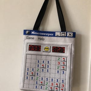 May include: A light grey tote bag with black shoulder straps featuring a printed graphic of the classic Minesweeper computer game interface. The design includes the title bar, a yellow smiley face icon, and a grid filled with colourful numbers.