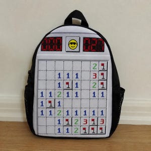 Peut inclure: Un sac à dos noir avec un panneau avant imprimé représentant l'interface du jeu informatique classique Démineur. Le motif comprend une grille de carrés numérotés, des icônes de drapeaux rouges, une barre d'état avec un smiley jaune et des poches latérales en filet noir.