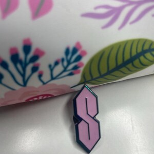 May include: A pink and blue enamel pin in the shape of the 'S' symbol, a popular design from the 1990s. The pin has a light pink interior and a dark blue outline. The background features a floral pattern with pink and purple flowers.