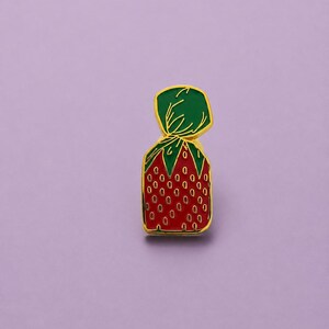 Peut inclure: Une broche en émail à bordure dorée en forme de bonbon à la fraise enveloppé dans un emballage vert torsadé. Le corps du bonbon est rouge avec un motif répétitif de petites graines ovales dorées, et la partie supérieure est d'un vert vif.