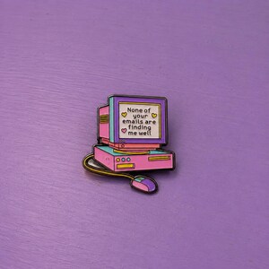Peut inclure: Une broche en émail métallique en forme d'ordinateur de bureau rétro avec une base rose et un écran violet. L'écran affiche le texte 'None of your emails are finding me well' entouré de petits cœurs jaunes et roses et une souris d'ordinateur.