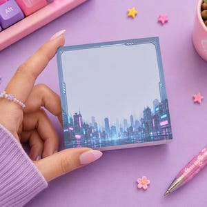 Peut inclure: Un bloc-notes carré autocollant orné d'une illustration imprimée d'un paysage urbain futuriste de nuit avec des néons bleus et roses. La note présente une bordure bleu foncé avec des accents géométriques et un centre gris clair vierge.