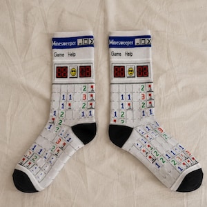 May include: A pair of white crew socks featuring a Minesweeper game interface print. The design includes a blue top bar with the text Minesweeper, Game, and Help, a smiley face icon, and a grid of colourful numbers and flags on a white background.