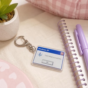 May include: A clear acrylic keychain with a Windows XP error message: "Task failed successfully." The keychain has a silver metal ring and clasp. A notebook, pen, and small potted plant are in the background.
