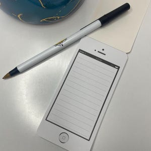 May include: A white smartphone-shaped notepad with a lined interior, a black pen, and a decorative ceramic container. The notepad has a screen display that reads "9:41 AM". The pen is white with a black tip.