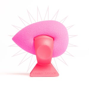 May include: A bright pink makeup sponge with a teardrop shape, resting on a matching pink stand. The stand has a flared base and the text "TAMMY TAYLOR" is printed on the base. The background features a subtle starburst design.