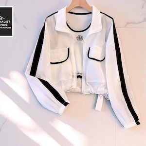 Peut inclure: Veste blanche avec des rayures noires sur les manches et une bordure noire. La veste a deux poches et un motif de rose sur la poitrine. Le texte "TEENS MINIMALIST CLOTHING UNCLUTTEREDWARDROBE" est dans le coin supérieur gauche.