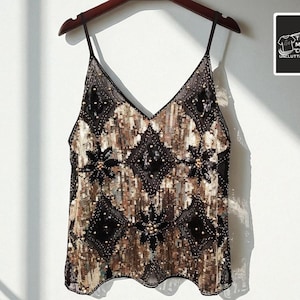 May include: A V-neck camisole top with a black and gold sequin design. The top features a geometric pattern with diamond shapes and floral accents. The garment hangs on a wooden hanger.
