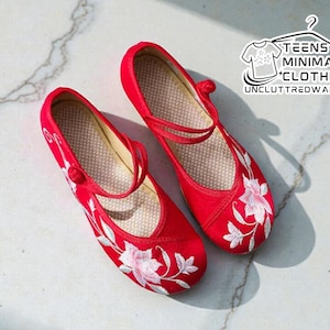 May include: A pair of red flats with white floral embroidery. The shoes have a strap across the top of the foot and a small button closure. The shoes are from the Teens Minimalist Clothing brand.