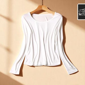 Pode incluir: Uma camisa branca de manga comprida com decote redondo está pendurada em um cabide de madeira. O tecido parece macio e esvoaçante. Um pequeno logotipo quadrado com texto está no canto superior direito.