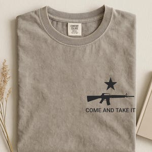 May include: A gray Comfort Colors t-shirt with a black graphic of a rifle and star, with the text "COME AND TAKE IT". The shirt is folded and displayed on a neutral surface.