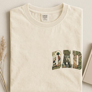 May include: Cream-coloured t-shirt featuring the word "DAD" in a nature-themed design. The letters incorporate images of a duck, binoculars, and a deer. The shirt has a crew neck and a small tag on the collar.