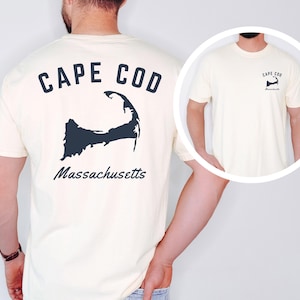 Puede incluir: Camiseta color crema con el texto "CAPE COD" sobre una silueta azul oscuro de Cape Cod, Massachusetts. La palabra "Massachusetts" está impresa debajo. Una versión más pequeña del diseño está en la parte delantera.